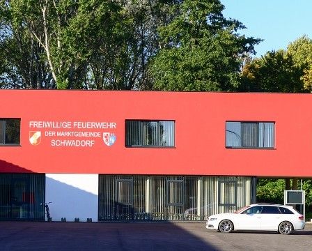 Ein rotes Gebäude mit weißem Rand und großen Fenstern steht in einem grünen Bereich. Ein weißes Auto ist davor geparkt. Das Schild steht für 'Freiwillige Feuerwehr der Marktgemeinde Schwadorf.' Straßenlaternen und Bäume im Hintergrund.