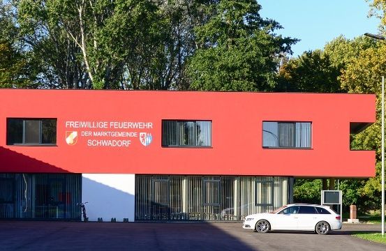 Ein rotes Gebäude mit weißem Rand und großen Fenstern steht in einem grünen Bereich. Ein weißes Auto ist davor geparkt. Das Schild steht für 'Freiwillige Feuerwehr der Marktgemeinde Schwadorf.' Straßenlaternen und Bäume im Hintergrund.