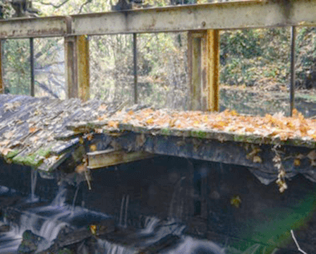 Ein Blick auf eine kleine Brücke über einem fließenden Bach, mit verstreuten gefallenen Blättern auf der Brücke und einem Wald im Hintergrund.