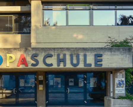 Der Eingang eines Gebäudes mit 'EUROPASCHULE' in bunten Buchstaben über den Eingangstüren. Das Gebäude hat Glasfenster und einen Schatten auf der Wand.