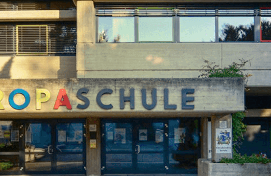 Der Eingang eines Gebäudes mit 'EUROPASCHULE' in bunten Buchstaben über den Eingangstüren. Das Gebäude hat Glasfenster und einen Schatten auf der Wand.