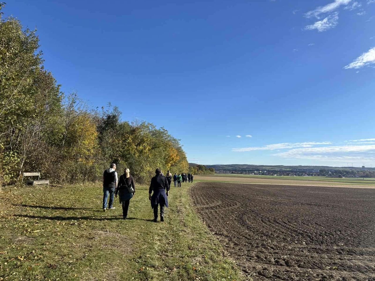 Eine Gruppe von Menschen geht entlang eines grasigen Pfades in der Nähe eines gepflügten Feldes. Der Himmel ist klar und blau.