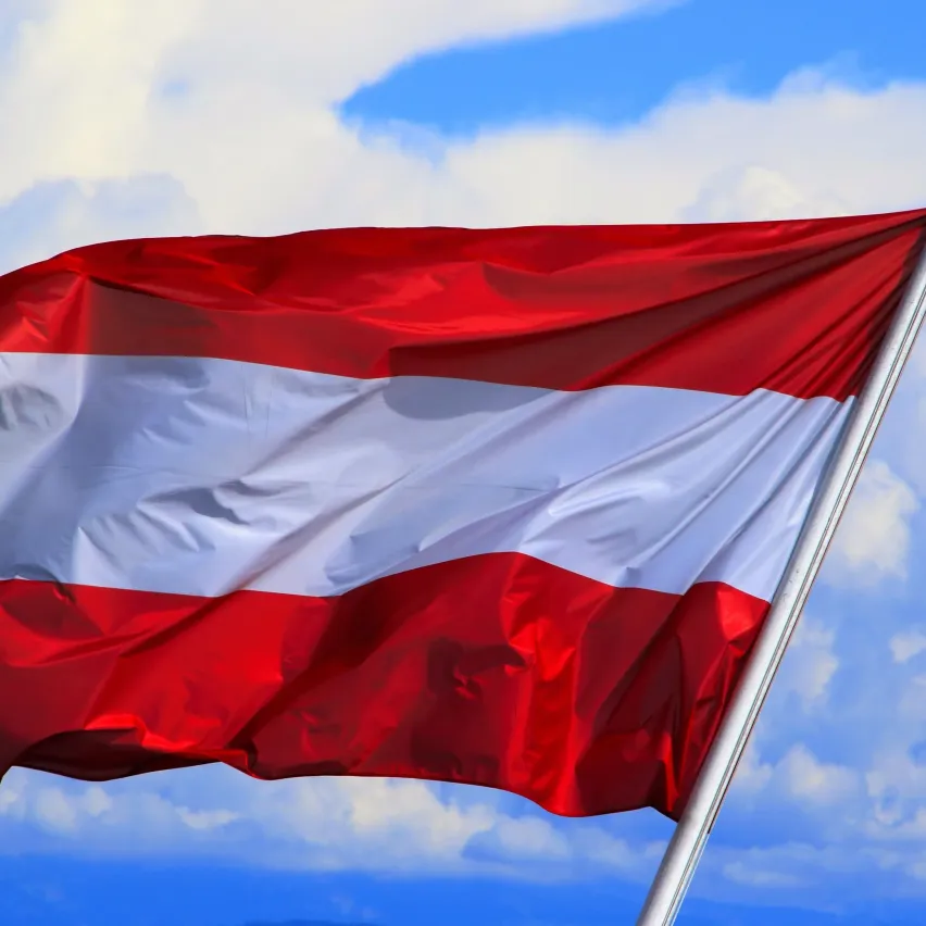 Eine rot-weiße Flagge mit einem zentralen Emblem, die im Wind gegen einen blauen Himmel mit Wolken weht.
