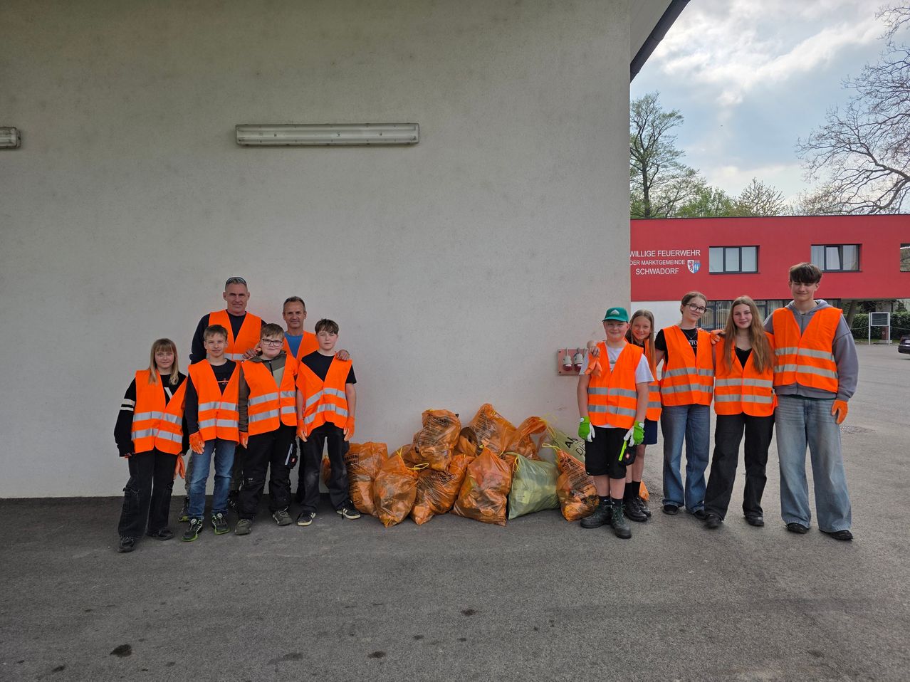 Eine Gruppe von Menschen in orangefarbenen Westen posiert vor einem Feuerwehrhaus. In der Nähe befinden sich Müllsäcke.