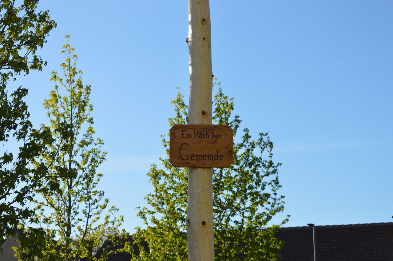 Ein Holzschild mit der Aufschrift 'Ein Hoch der Gemeinde' ist an einem Holzpfahl zwischen Bäumen montiert. Der Himmel ist klar und blau.