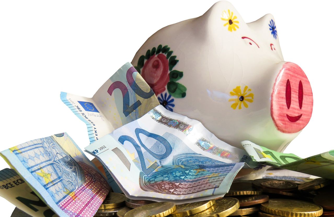 Ein Nahaufnahme eines Sparschweins mit europäischer Währung vor weißem Hintergrund. Das Schwein hat Blumen und Rosen gemalt. Mehrere 20-Euro-Scheine und Münzen sind darum verstreut.