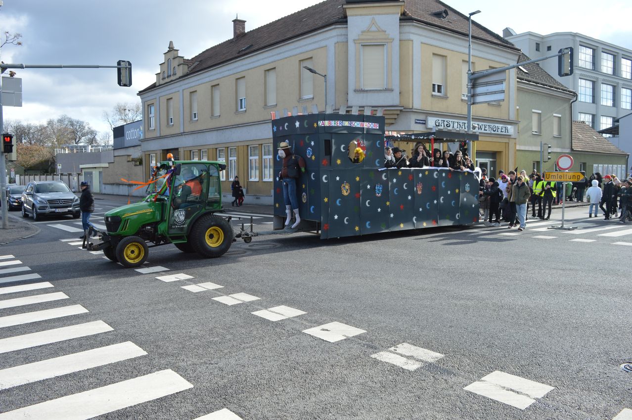Ein Umzug mit einem geschmückten Traktor, der einen Anhänger mit Personen darin zieht, vorbei an einem Gebäude mit dem Schild 'Gasthaus Weber'.