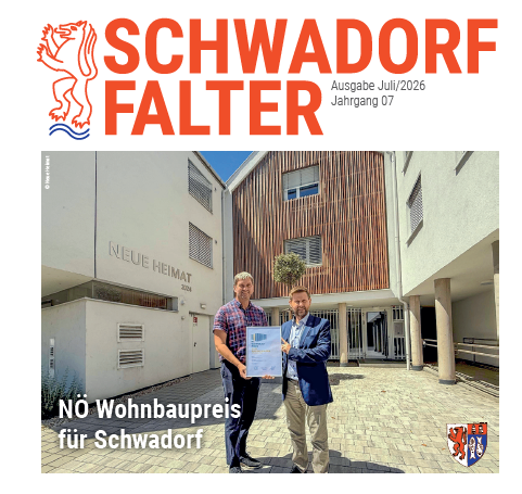 Zwei Männer stehen vor einem Gebäude mit einem Schild, das 'NÖ Wohnbaupreis für Schwadorf' besagt. Der Mann auf der linken Seite hält ein Zertifikat.