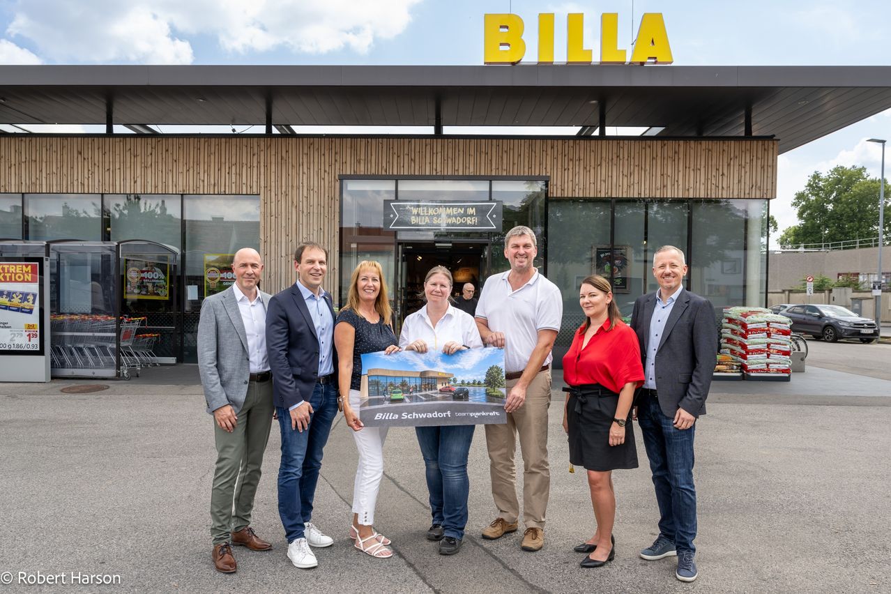 Eine Gruppe von Menschen steht vor einem BILLA-Geschäft und hält ein Schild mit der Aufschrift BILLA Schwadorf. Sie alle lächeln und scheinen für ein Foto zu posieren.