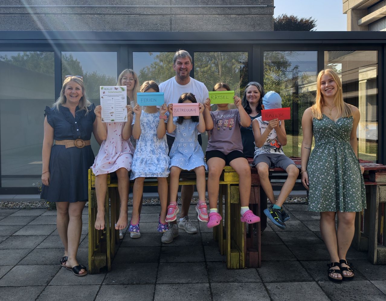 Eine Gruppe von Menschen, einschließlich Kindern, posiert für ein Foto und hält bunte Karten mit darauf geschriebenen Wörtern. Sie stehen auf einer Holzbank vor einem Gebäude.