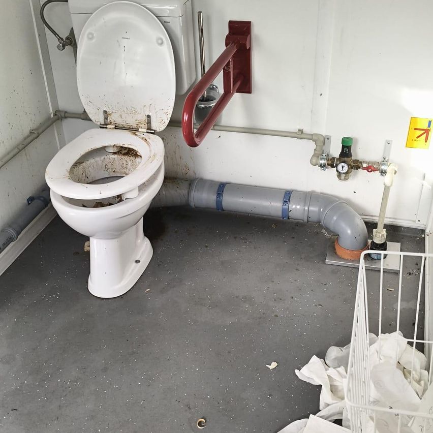 Eine schmutzige Toilette mit offenem Deckel in einem kleinen Raum mit Rohren und einem weißen Korb in der Nähe.