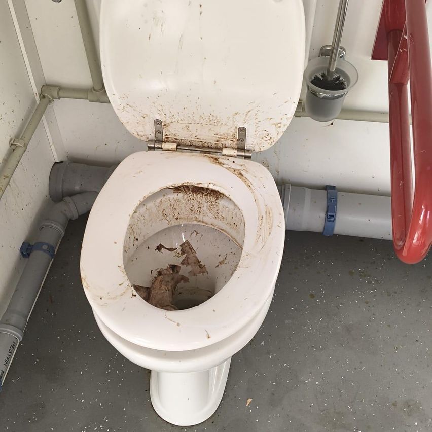 Eine schmutzige weiße Toilette mit offenem Deckel, gefüllt mit braunem Wasser und Ablagerungen, in einem Badezimmer mit sichtbaren Rohren und einem roten Griff.