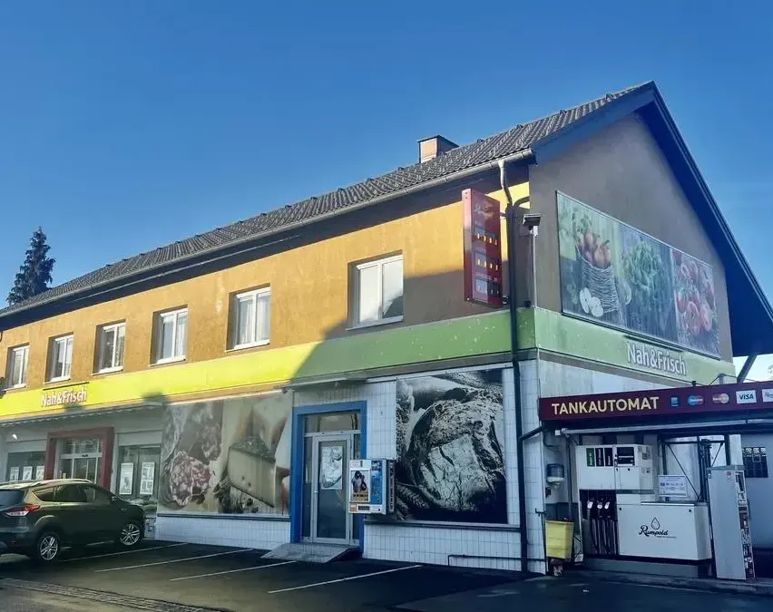 Ein Geschäft mit einer gelben und grünen Fassade und einem Schild, das 'Nah&Frisch' steht, befindet sich an einer Straße. Das Geschäft hat ein großes Fenster mit einem Bild von Brot, Käse und Fleisch. Ein Zapfsäule befindet sich vor dem Geschäft. Ein Auto ist vor dem Geschäft geparkt.