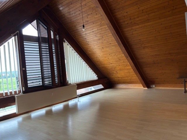 Ein leeres Dachzimmer mit Holzböden und -wänden, große Fenster mit Jalousien, eine Decke mit Holzbalken und eine hängende Lampe.