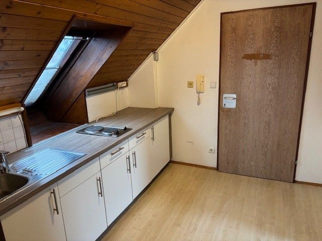 Eine leere Küche mit Holzboden, einer Holztür und einem Waschbecken mit Wasserhahn. Sie hat einen Gasherd, Schränke und ein an der Wand montiertes Telefon.