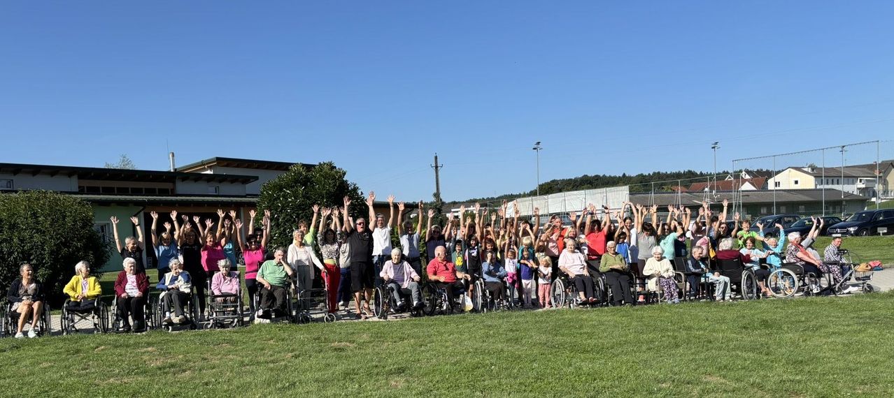Eine Gruppe von Menschen, einige in Rollstühlen, posiert für ein Foto an einem sonnigen Tag und hebt die Hände. Hinter ihnen sind ein Gebäude, Bäume und Straßenlaternen zu sehen.