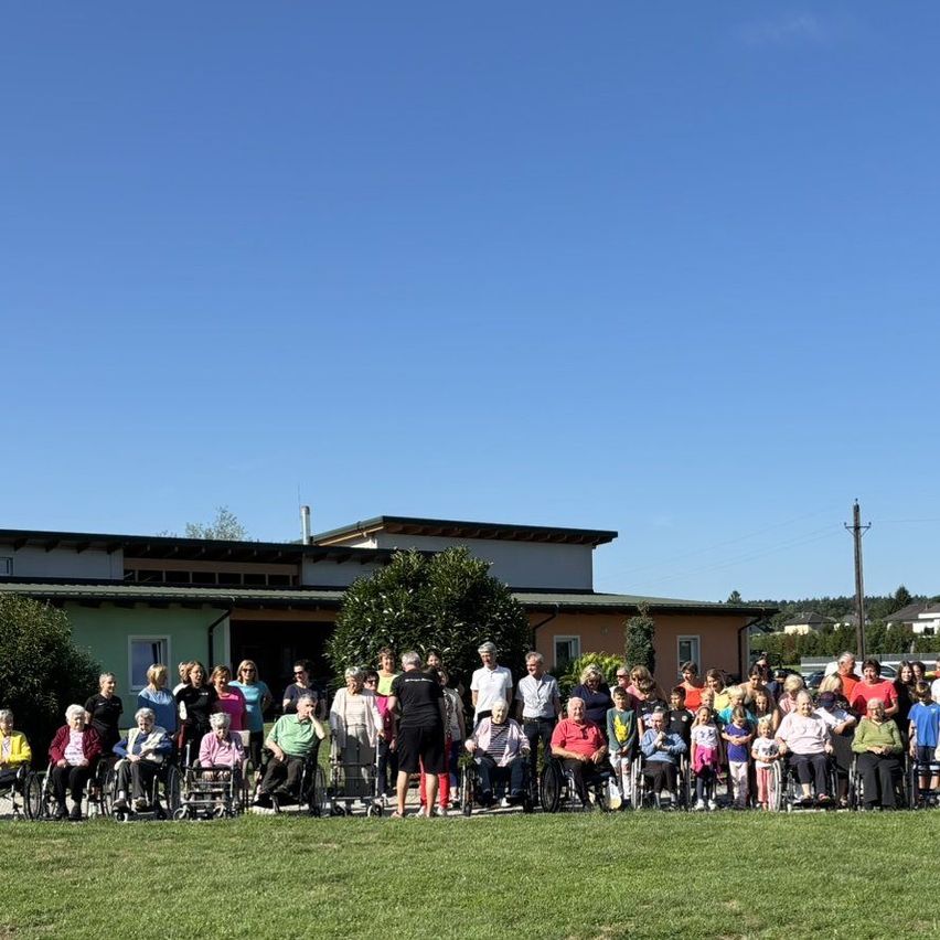 Eine Gruppe älterer Menschen in Rollstühlen posiert für ein Foto vor einem Gebäude mit klarem blauem Himmel.