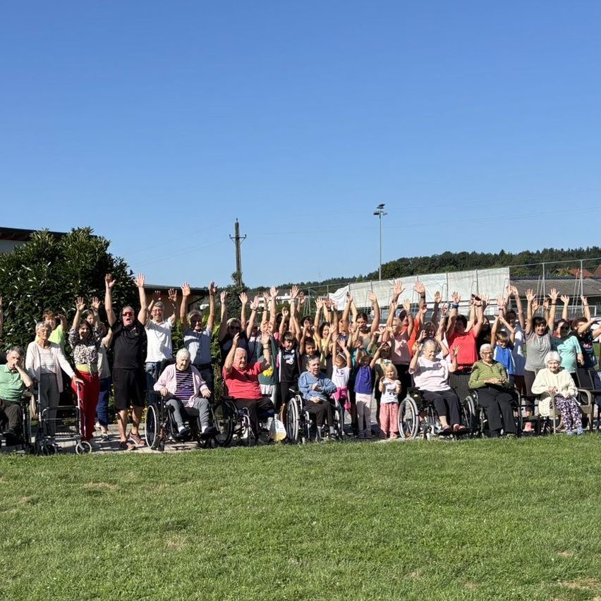 Eine Gruppe von Menschen, darunter einige in Rollstühlen, versammelt sich auf einem Rasenfeld. Sie heben alle ihre Hände in die Luft. Bäume, Gebäude und Straßenlaternen sind im Hintergrund zu sehen.