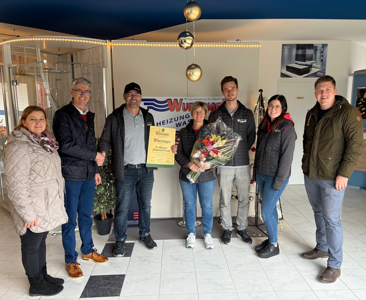 Eine Gruppe von Menschen steht in einem Raum, lächelt und posiert für ein Foto. Eine Frau hält einen Blumenstrauß. Ein Mann hält ein Zertifikat und steht neben einer Frau. Hinter ihnen hat eine weiße Wand ein Banner mit dem Text 'Würzner'.