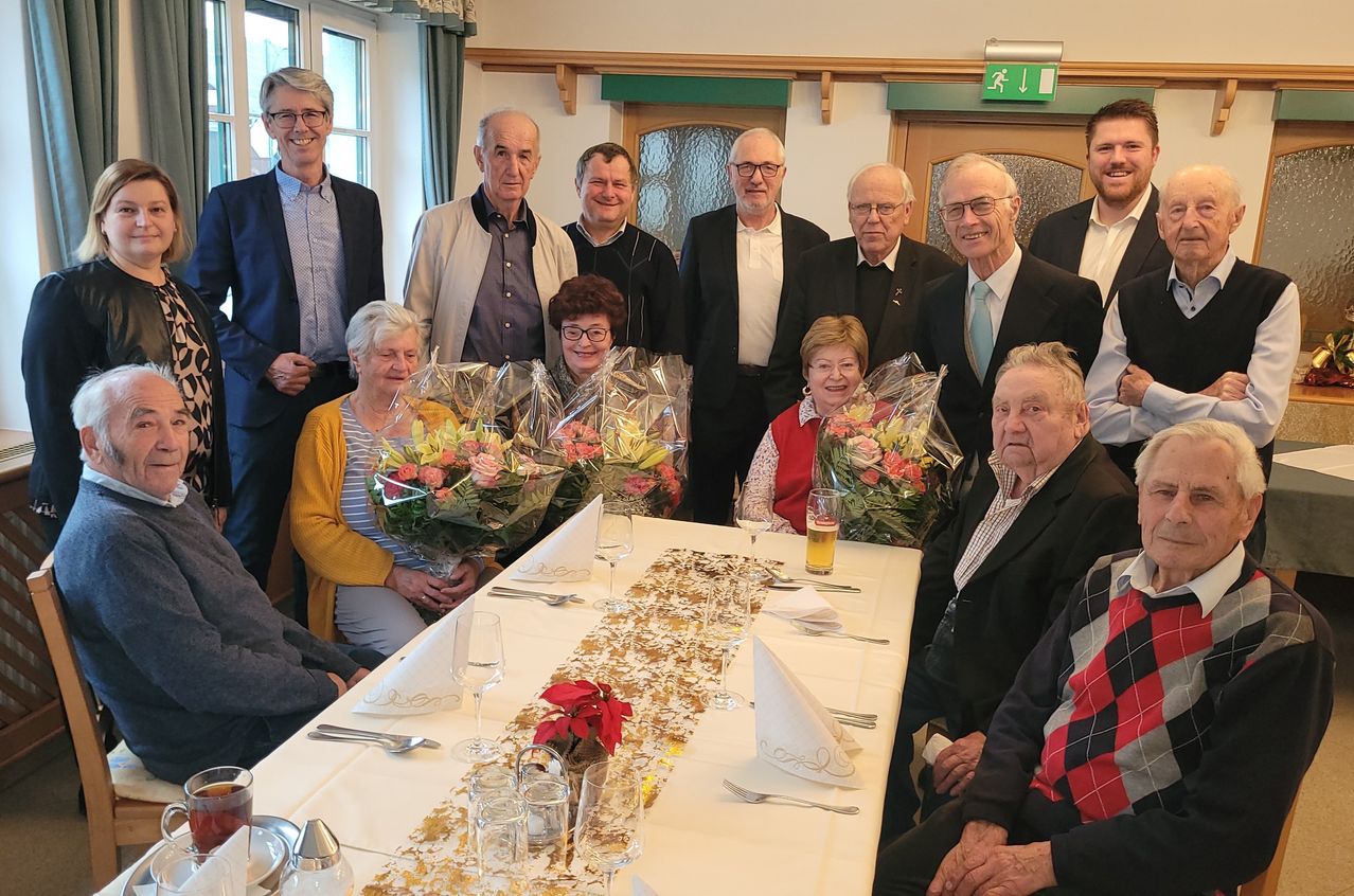 Eine Gruppe von Menschen versammelt sich um einen Tisch, lächelt und posiert für ein Foto. Blumen und Getränke stehen auf dem Tisch, mit einem Fenster und einer Tür im Hintergrund.