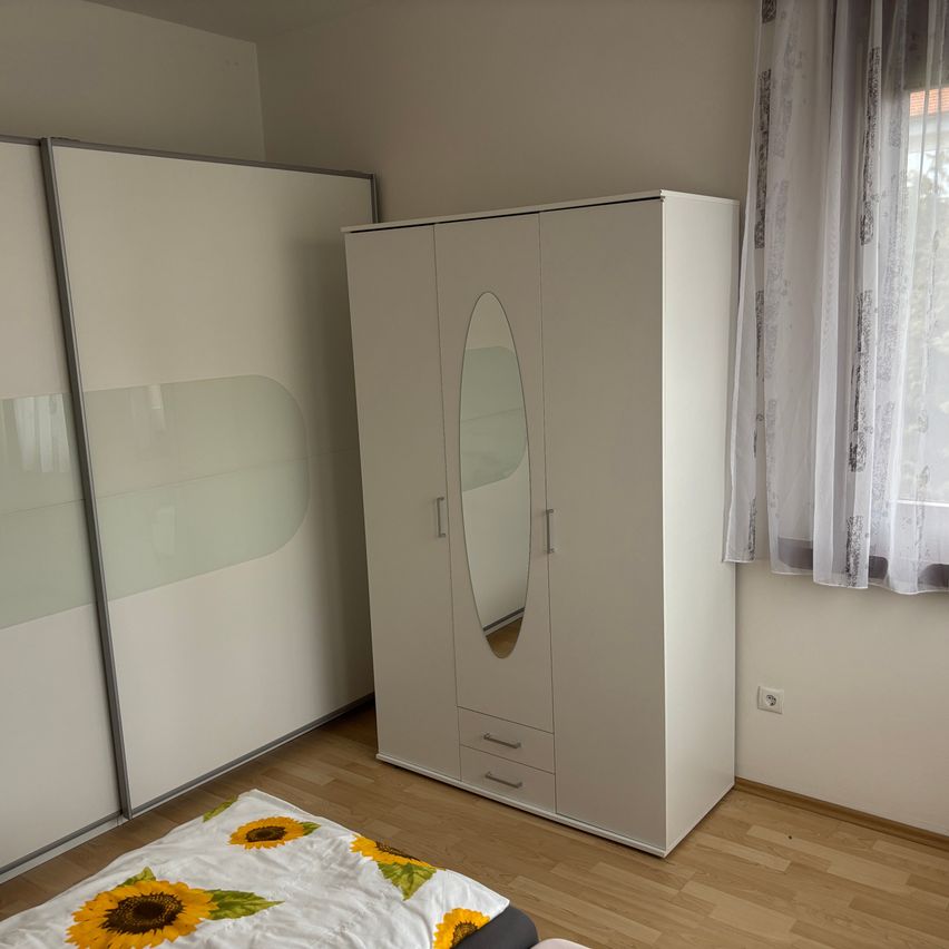 Ein Schlafzimmer mit einem weißen Kleiderschrank mit Spiegel und zwei Schubladen. Ein Bett mit einer sonnenblumenbedruckten Decke im Vordergrund. Der Raum hat eine weiße Wand und eine weiße Gardine in der Nähe eines Fensters.