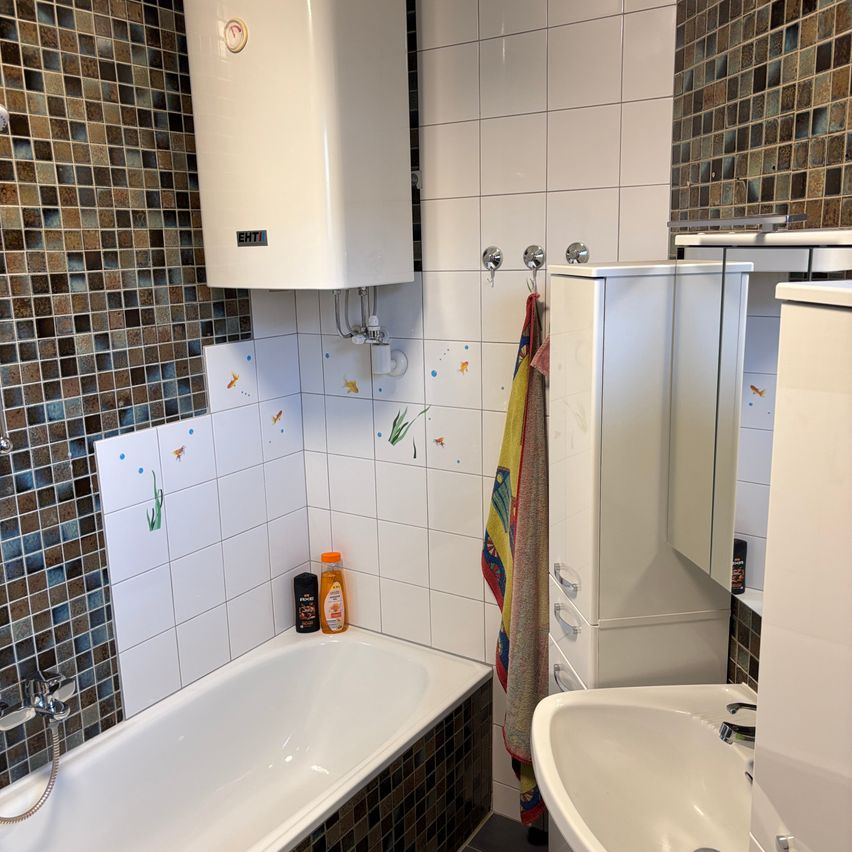 Ein Badezimmer mit Badewanne, Waschbecken und Schrank. Die Wände sind gefliest, und ein Handtuch hängt an einem Haken.