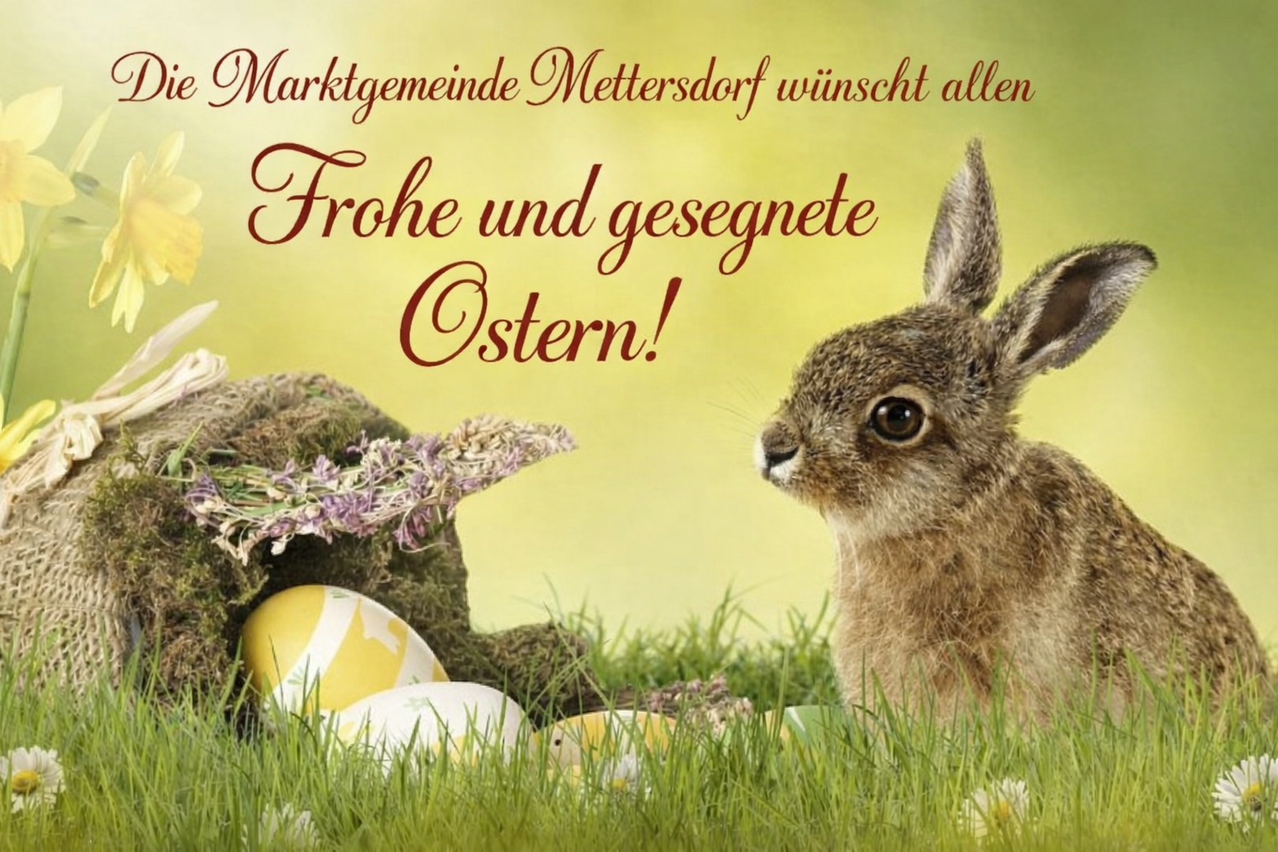 Ein Kaninchen mit braunem Fell sitzt auf dem Gras in der Nähe von Ostereiern. Ein Banner darüber lautet 'Marktgemeinde Mettersdorf wünscht allen ein frohes und gesegnetes Ostern!'.