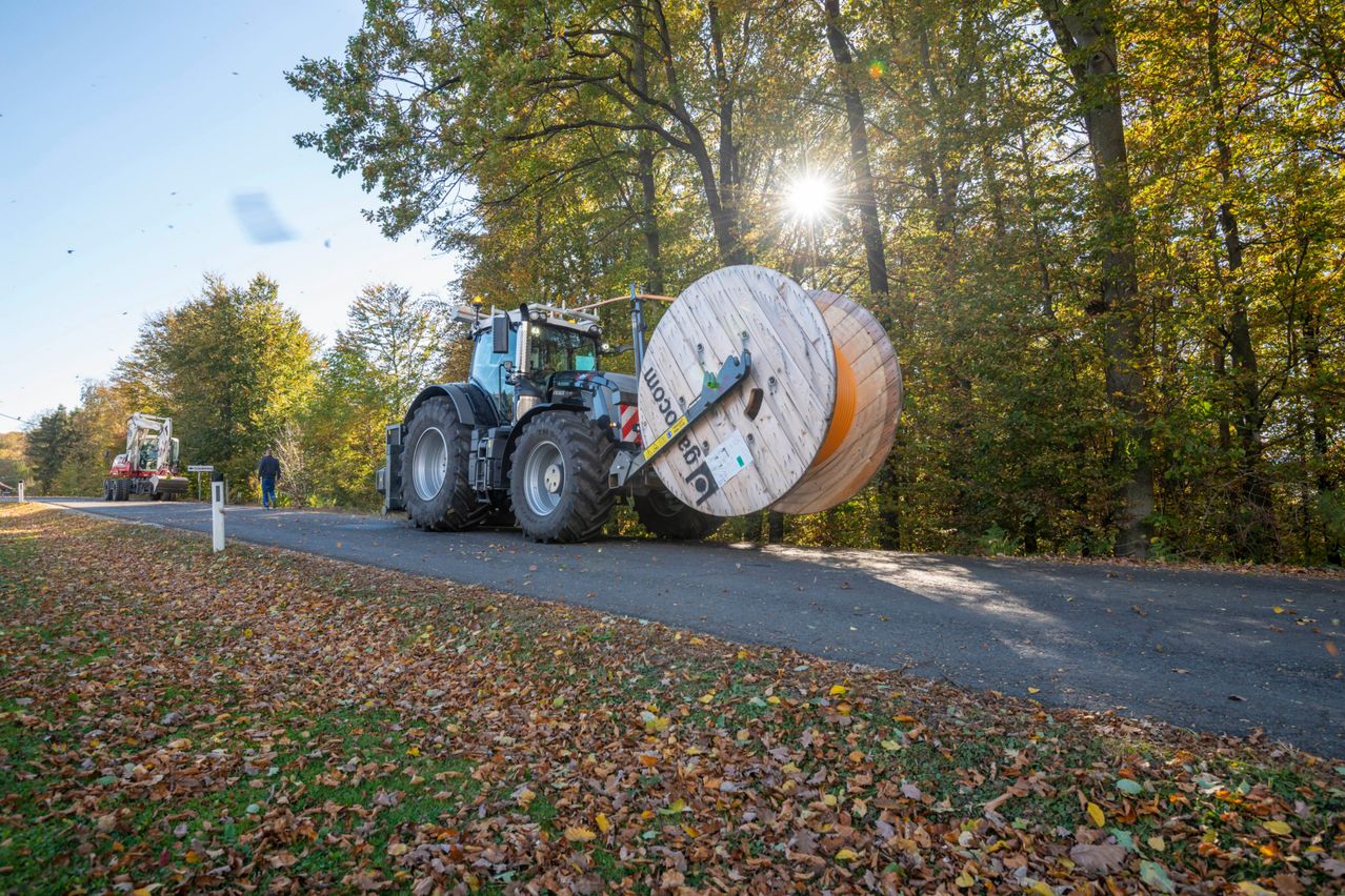 A tractor with large wooden spools attached is driving on a road surrounded by trees in autumn. A person is walking nearby.
