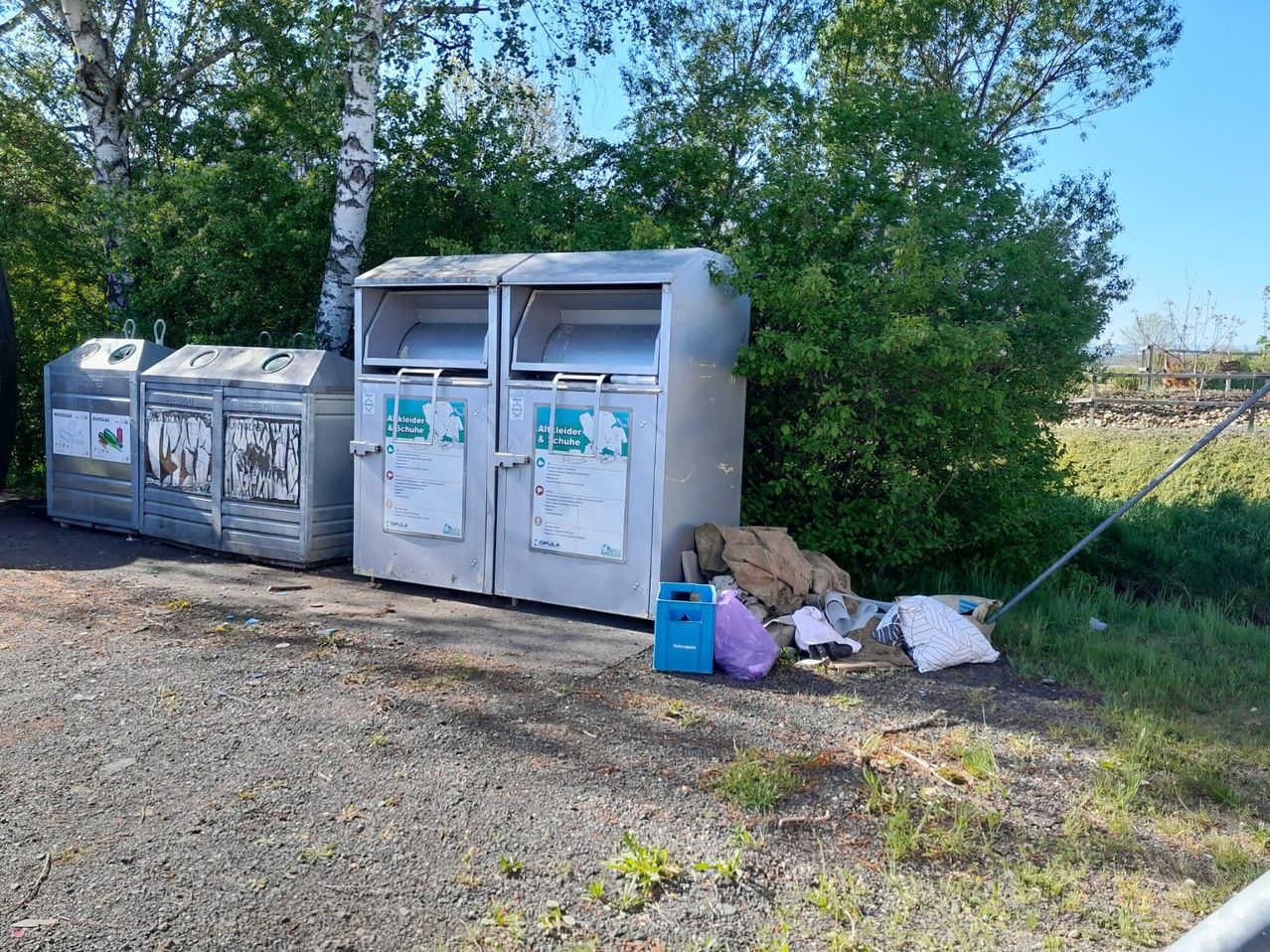 Several metal trash bins are on a gravel lot. One bin is open, and waste is spilled out. Trees are behind the bins.