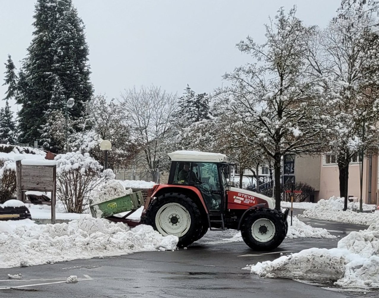 A red and white tractor is plowing snow on a road with a green plow attached, surrounded by snow-covered trees and bushes.