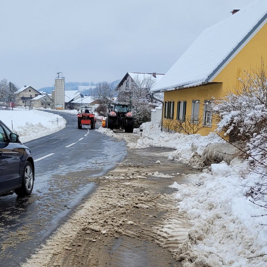A snowy road with a black car and two tractors. The road is wet, and there is a yellow house covered in snow.