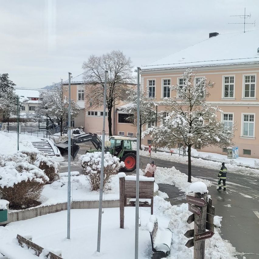 A snowy street with a green tractor and a person standing in the snow. A building with several windows and a roof covered in snow. Snow-covered trees and bushes are also present.