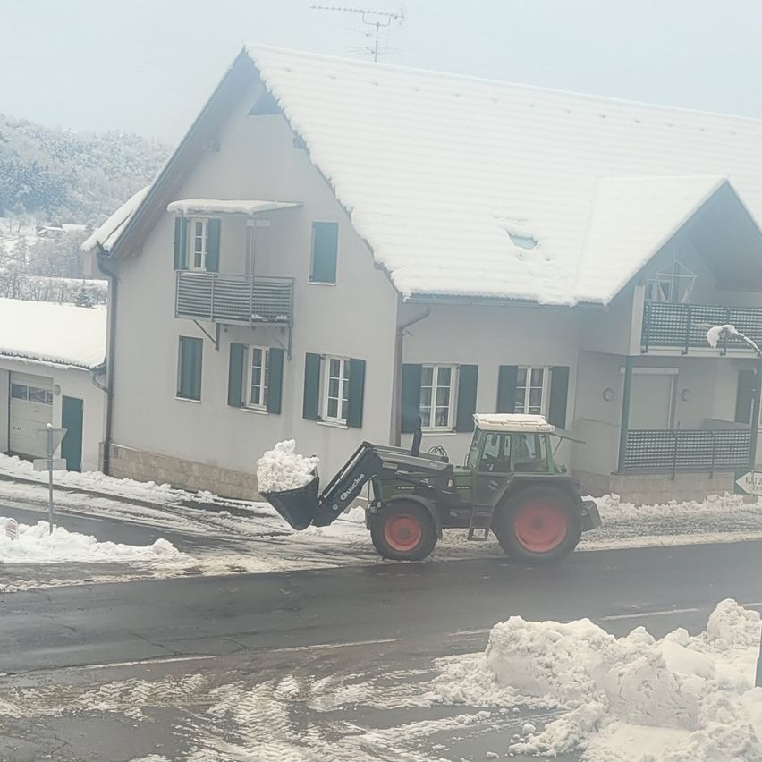 A tractor with a full bucket of snow is clearing a path in front of a house with snow on the roof and balconies.