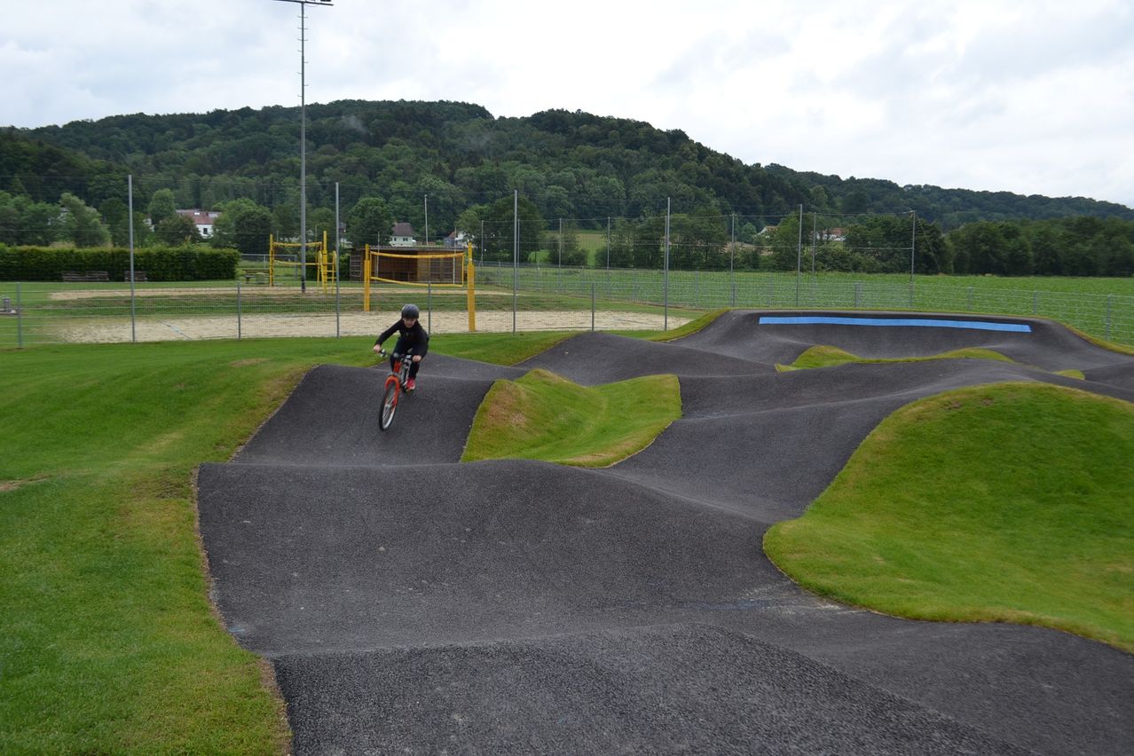 Ein Radfahrer mit Helm fährt einen kurvigen Pfad in einem Park hinunter, umgeben von Gras und Hügeln, mit einem Zaun und Spielplatz im Hintergrund.