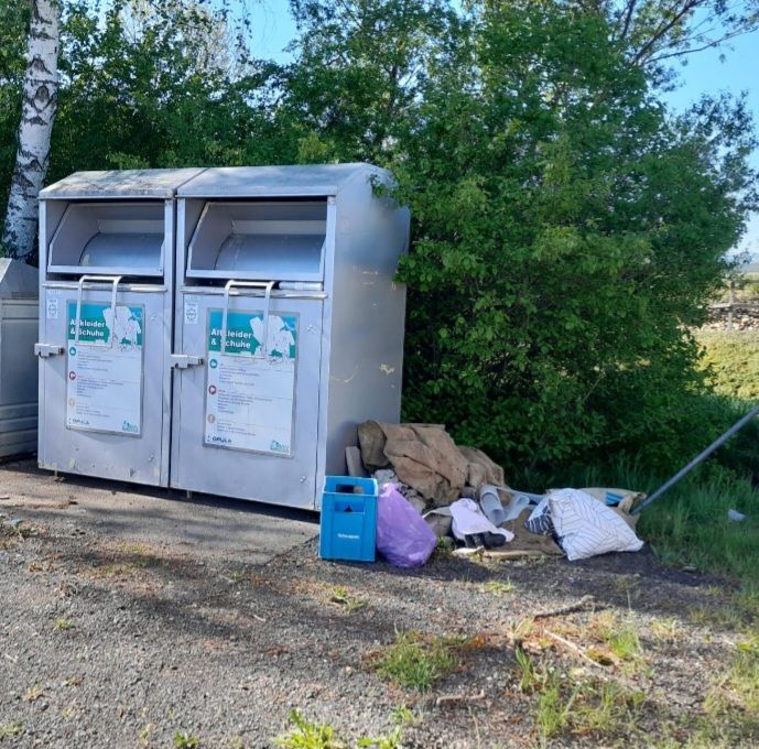 Zwei Metallrecyclingbehälter mit deutschen Anweisungen stehen auf einem Schotterboden. In der Nähe befinden sich verstreute Gegenstände, darunter ein blauer Plastikbehälter und ein lila Beutel.