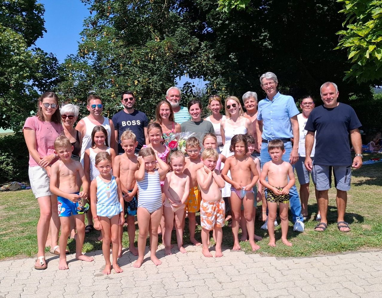 Eine Gruppe von Menschen, einschließlich Kindern und Erwachsenen, steht zusammen in einem Park und lächelt für ein Foto. Einige tragen Badekleidung.