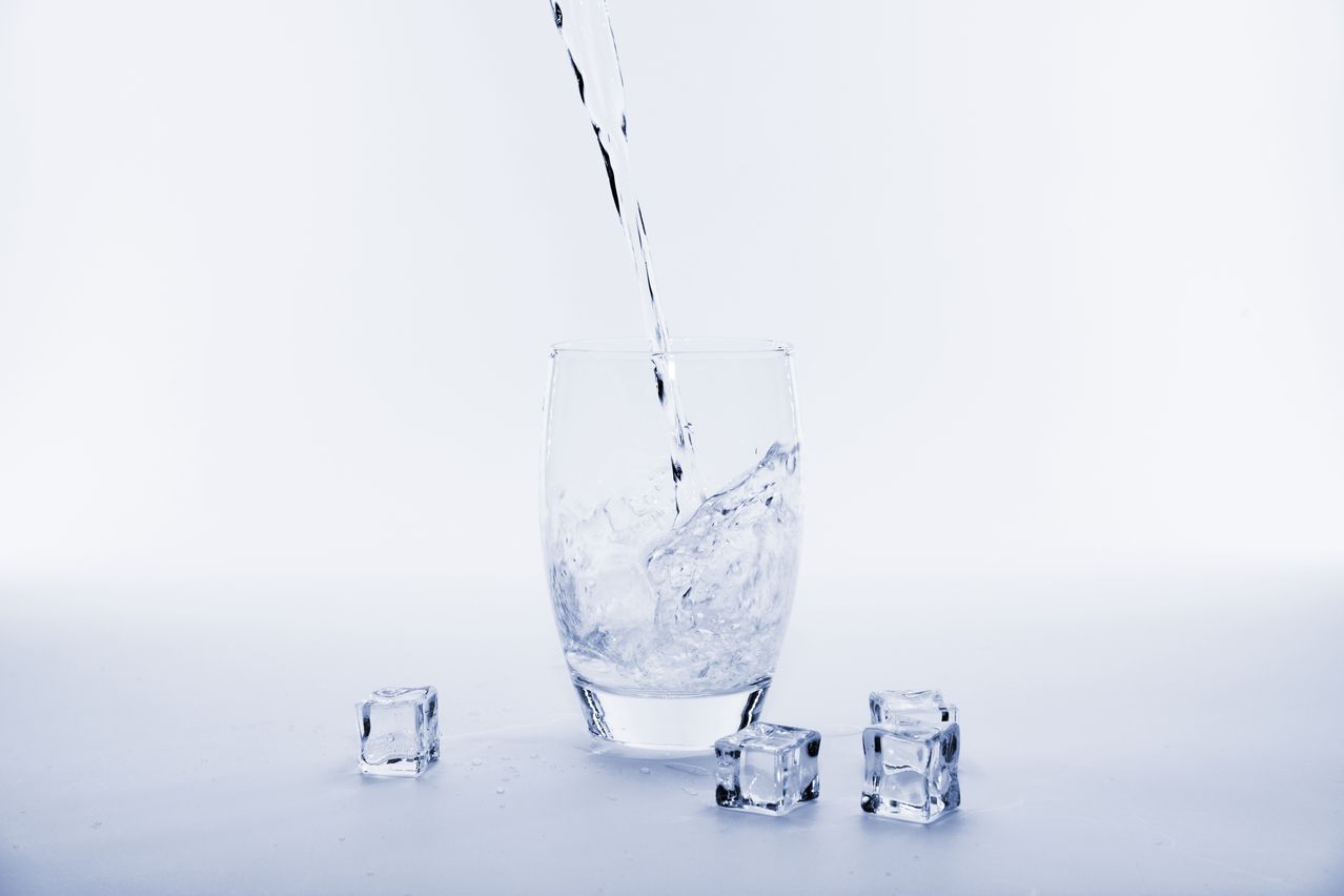 Ein klares Glas mit Wasser und Eiswürfeln, wobei Wasser von oben hineingegossen wird.