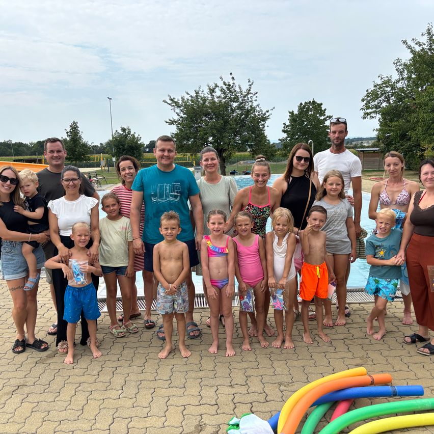 Eine Gruppe von Erwachsenen und Kindern posiert für ein Foto vor einem Schwimmbad mit bunten Poolspielzeug.