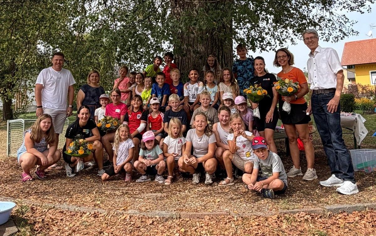 Eine Gruppe von Kindern und Erwachsenen posiert für ein Foto unter einem großen Baum, einige halten Blumen.