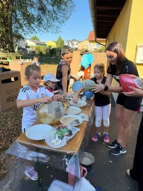 Eine Gruppe von Kindern und Erwachsenen versammelt sich um einen Tisch mit Tellern, Schüsseln und Luftballons. Die Kinder sind in einer Aktivität verwickelt, möglicherweise Basteln oder Dekorieren. Die Erwachsenen helfen ihnen. Die Umgebung scheint draußen zu sein, mit einem Gebäude und Bäumen im Hintergrund.