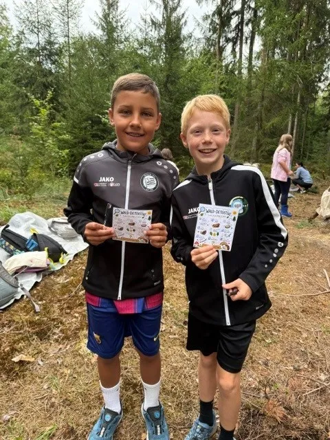 Zwei junge Jungen in passenden schwarzen Jacken mit weißen Streifen stehen in einem Wald und halten Karten mit Zeichnungen in der Hand.