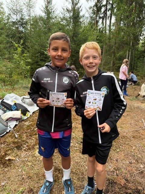 Zwei junge Jungen in passenden schwarzen Jacken mit weißen Streifen stehen in einem Wald und halten Karten mit Zeichnungen in der Hand.
