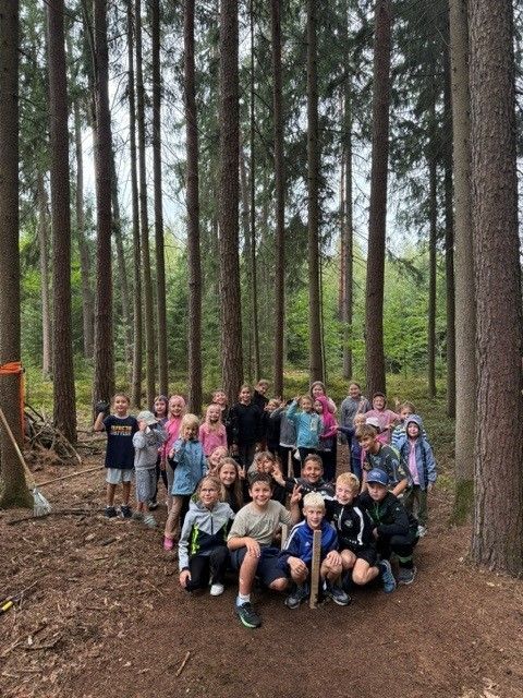 Eine Gruppe von Kindern und Erwachsenen posiert für ein Foto im Wald, einige halten Stöcke und andere stehen im Hintergrund.
