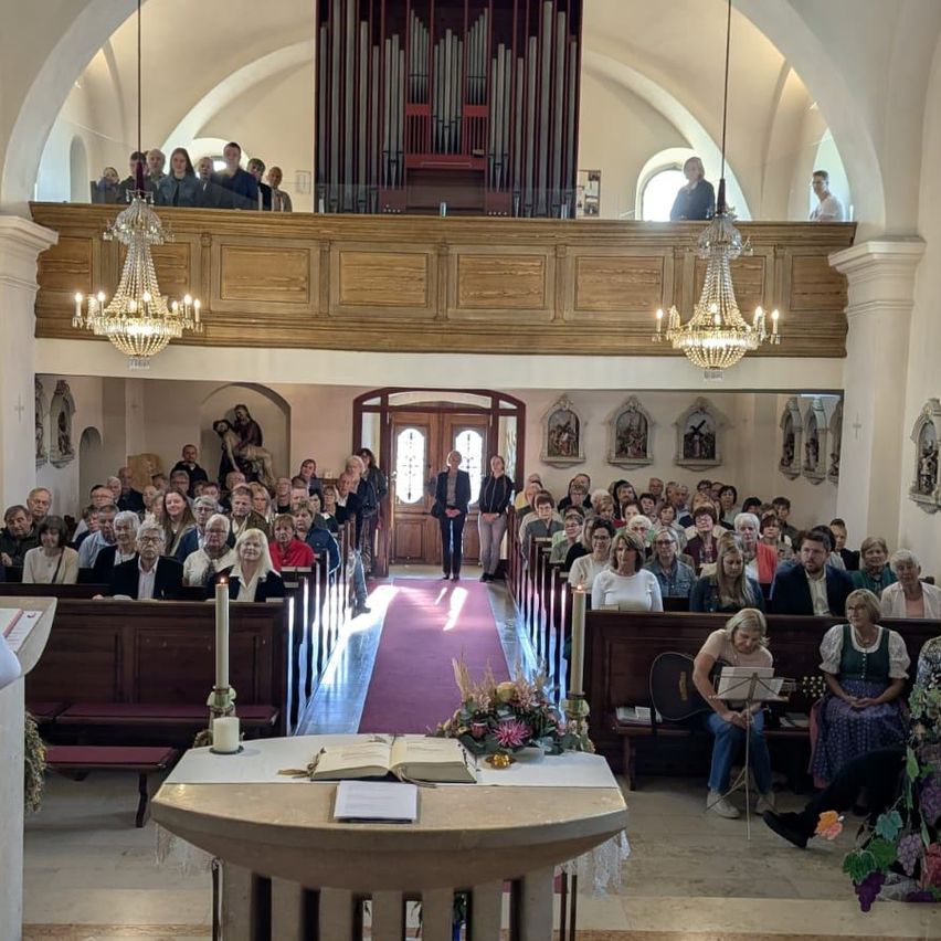 Eine große Gemeinde versammelt sich in einer Kirche, mit einem Chor auf der Empore und einer Person, die Gitarre spielt im Vordergrund. Kerzen und Blumen schmücken den Altar.