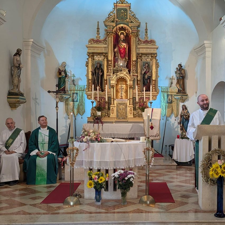 Ein Priester steht am Altar in einer Kirche, während zwei andere Priester in der Nähe sitzen. Der Altar ist mit Blumen, Kerzen und einer Statue von Jesus Christus geschmückt. Die Kirchenwände sind mit Statuen und Gemälden verziert.