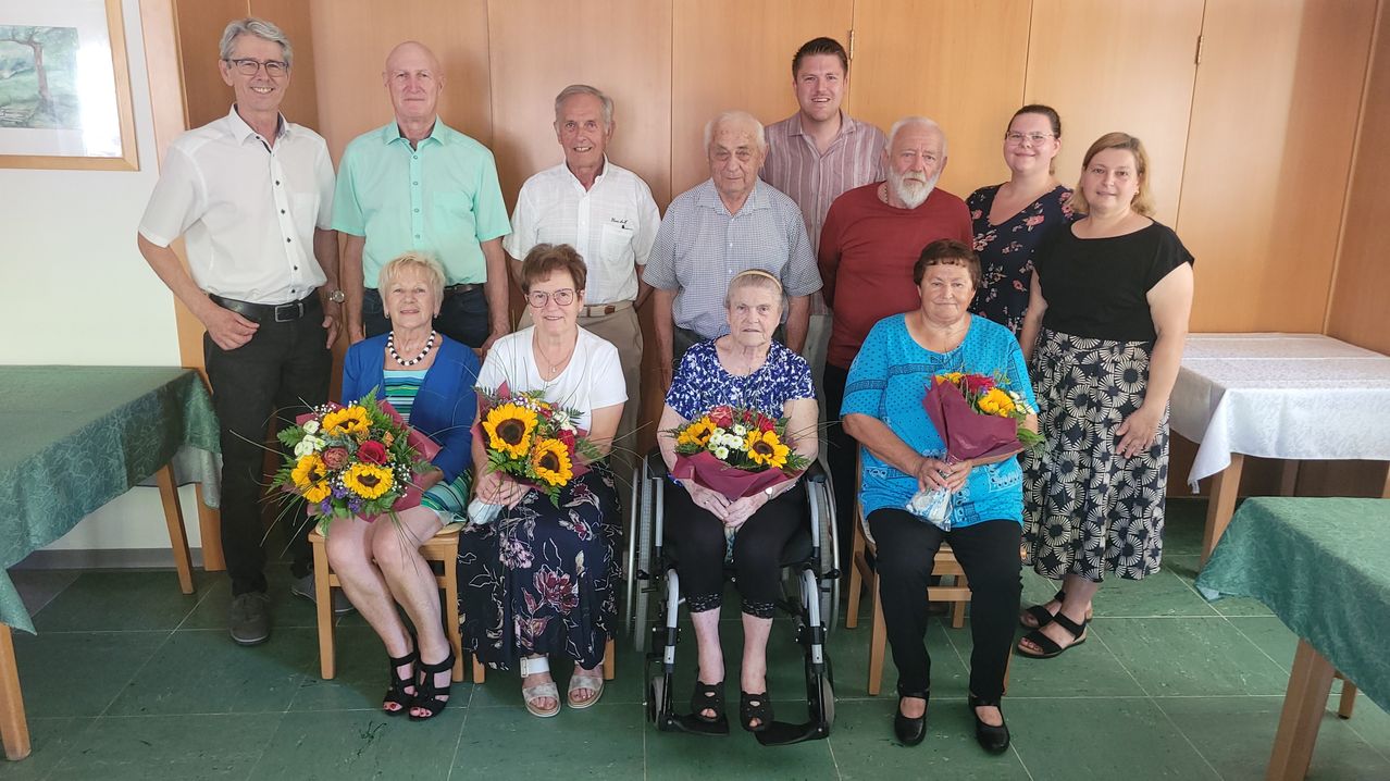 Eine Gruppe älterer Menschen und einige Erwachsene versammeln sich, einige in Rollstühlen, und halten Blumensträuße. Sie scheinen für ein Foto zu posieren.