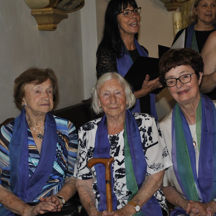 Drei ältere Frauen sitzen in einer Kirche zusammen. Eine hält einen Gehstock, eine andere ein Buch und die dritte trägt eine Halskette. Hinter ihnen stehen zwei Frauen, eine lächelt und hält ein Buch.