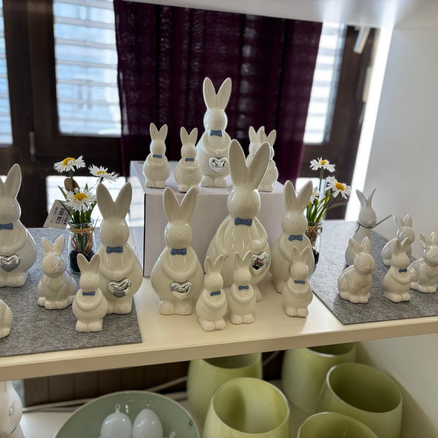 A display of white ceramic rabbits with blue bows and silver hearts is arranged on a white shelf. The rabbits are standing on a gray mat. A vase with daisies is placed on the left side. There are also some green ceramic vases on the bottom shelf.