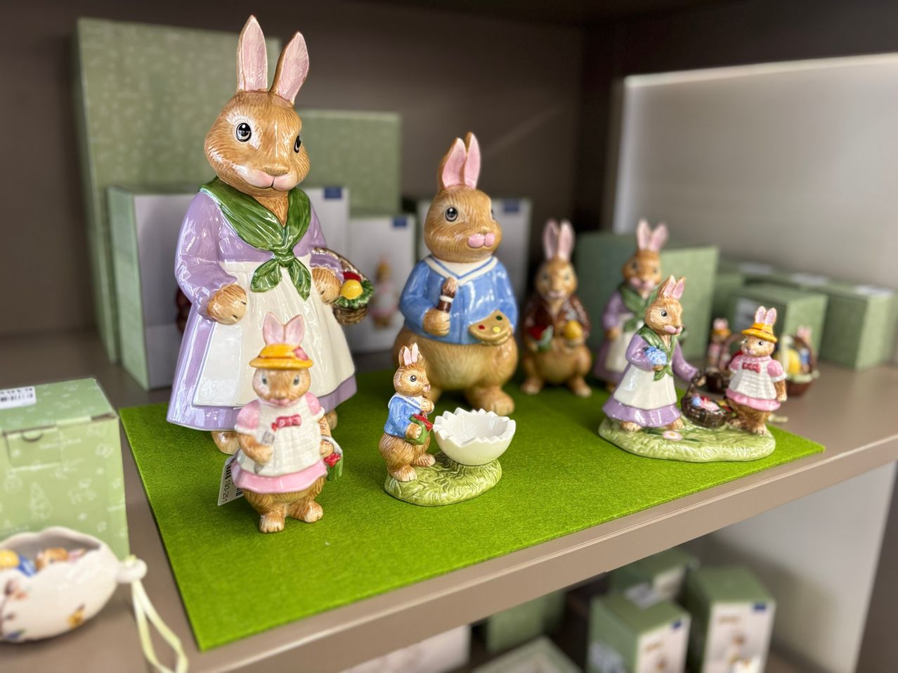A collection of Easter figurines of bunnies on a green mat, surrounded by boxes and other figurines. The main bunny holds a basket and wears a green scarf.