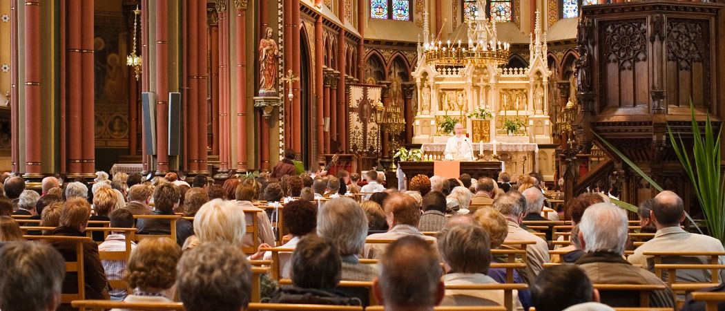 Ein Priester steht an einem Altar in einer Kirche mit Buntglasfenstern, einer Kronleuchter und einer Statue. Viele Menschen sitzen auf Kirchenbänken und hören aufmerksam zu.